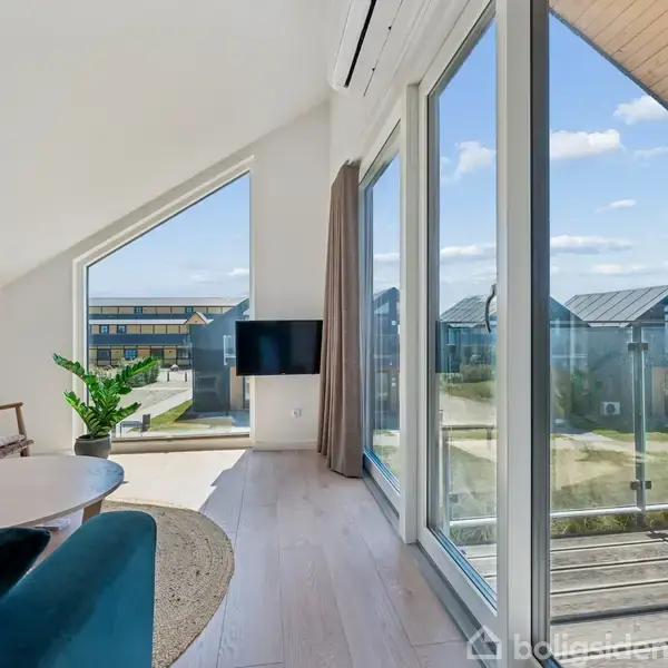 Stue med sofa og sofabord ved siden af store vinduer, der giver udsigt til moderne boligbyggeri udenfor. En fladskærm hænger på væggen, og der står en plante.