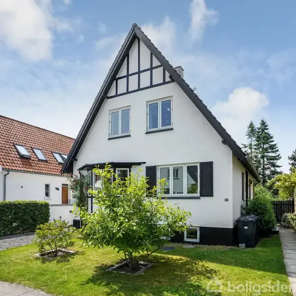 Et hvidt, to-etagers hus med skråt tag står i en velplejet have med grønne planter. Det er omgivet af andre huse i et beboelseskvarter.