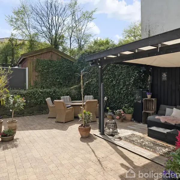 Terrasse viser en sofa under overdækning til højre, omgivet af planter i krukker. I baggrunden er der et udendørs siddesæt mod en grøn hæk og et træhus. Himlen er klar.