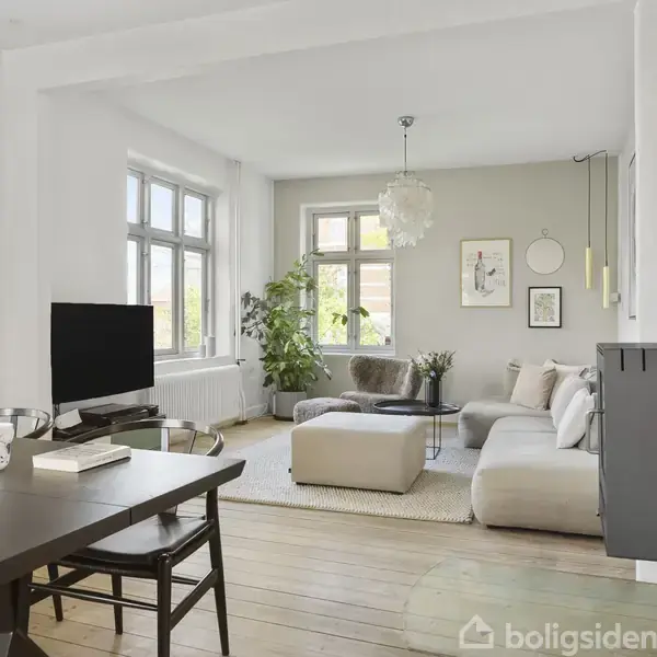 En lys stue med en stor grå sofa og sofabord, placeret på et trægulv. Planter og kunst på væggene skaber en hyggelig atmosfære. Fladskærms-TV ved siden af spisebord.