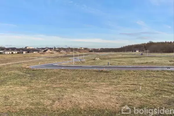 Græsplæne ligger stille i et åbent område med spredte huse i baggrunden. Asfalteret vej snor sig gennem landskabet under en klar, blå himmel.
