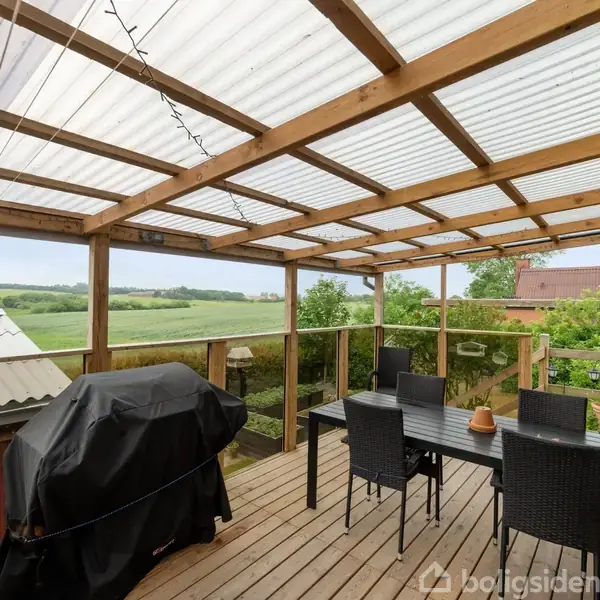 En overdækket terrasse med trægulv og gennemsigtigt tag. Der er et bord med seks sorte stole, en grill dækket med sort betræk, omgivet af grønne marker og en bygning.