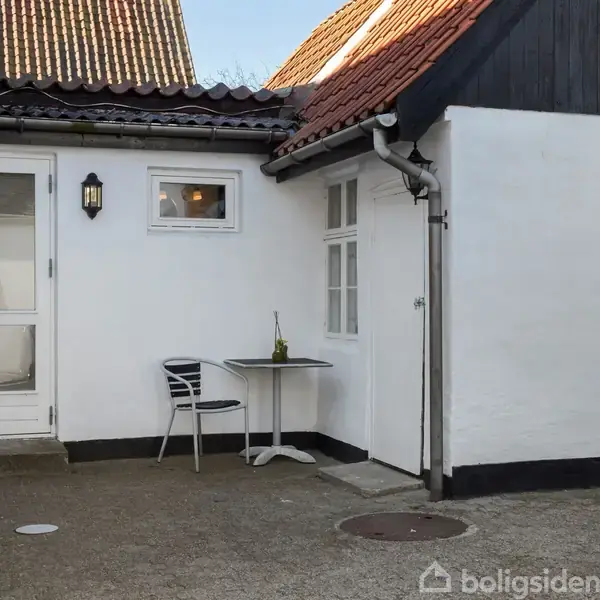 En lille gårdhave med et bord og en stol placeret mod en hvid væg. Der er en flaske som dekoration på bordet. Taget er skråt og dækket af tegl.