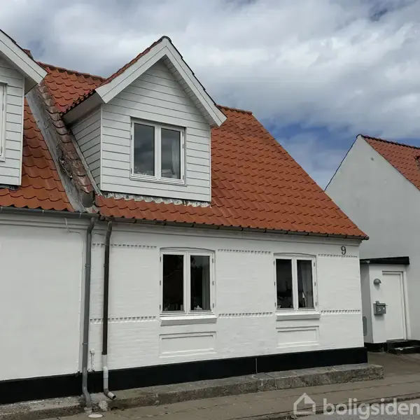 Et hvidt hus med rødt tegltag har en kvist med to vinduer. Et lille "9" er på facaden. Skyer fylder himlen i baggrunden.