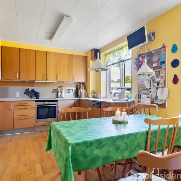 Et køkkenbord med grøn dug står dækket med lys, omgivet af træstole. Køkkenet har gule vægge, træskabe og to vinduer, der giver naturligt lys. En opslagstavle hænger ved siden af vinduerne.