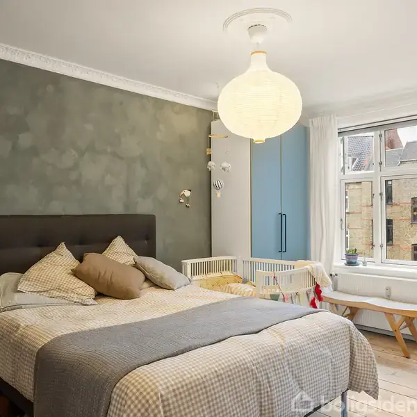 En dobbeltseng står centreret i et soveværelse med grøn accentvæg; den er dækket med flere puder og et tæppe. Et stort vindue og en himmelblå garderobe ses i baggrunden.