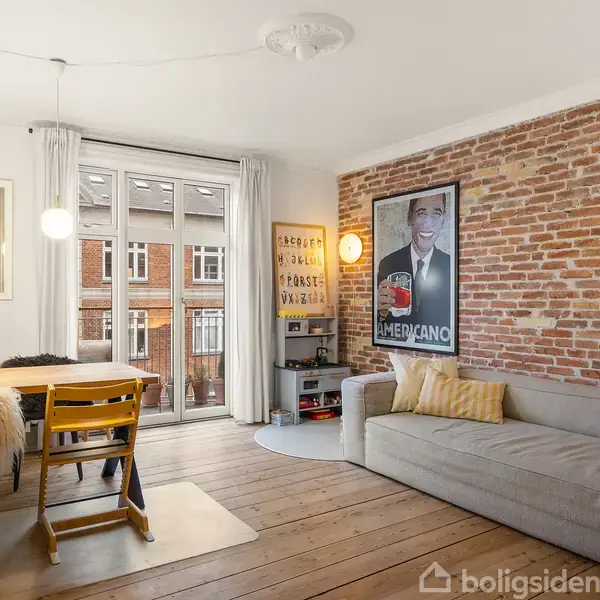 En stue med en grå sofa mod en murstensvæg; en plakat hænger over sofaen. Et spisebord med stole står nær en fransk altandør. En lampe hænger over bordet.