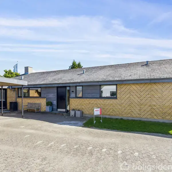 En gul murstensvilla med fladt tag og store vinduer står på en flisebelagt indkørsel. En cykel er parkeret ved carporten, og en græsplæne omkranser huset.