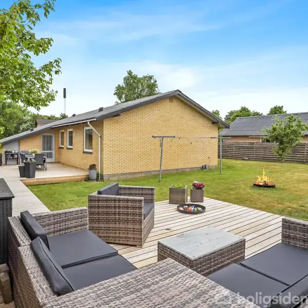Terrasse med grå flettemøbler, placeret på en træplatform. En gul murstensbygning ses i baggrunden med et udendørs spiseområde. Græsplæne og træer omgiver området.