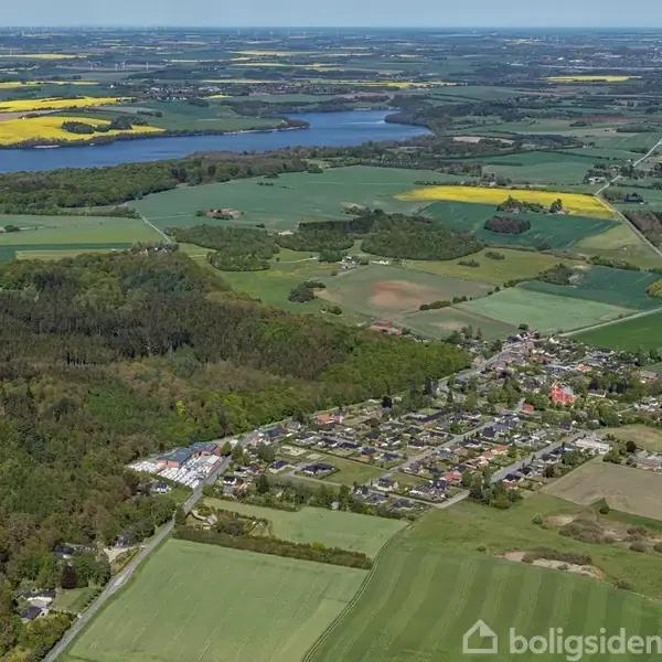 Landsby omgivet af marker og skove set fra luften, nær en flod. Området er præget af grønne marker, gule rapsmarker og skovområder spredt i landskabet.