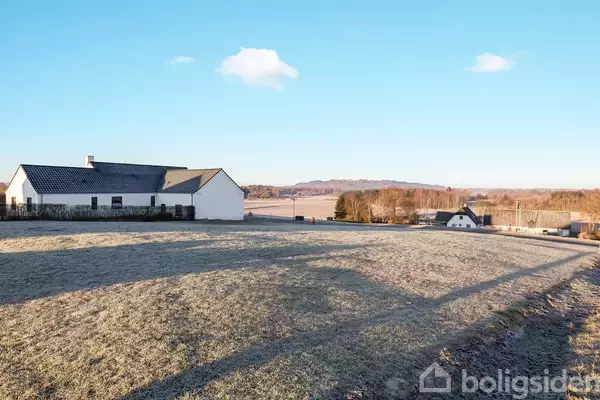 Hus står i solbeskinnet landskab med frostklædt græs og udsigt til åbne marker og træer i det fjerne.