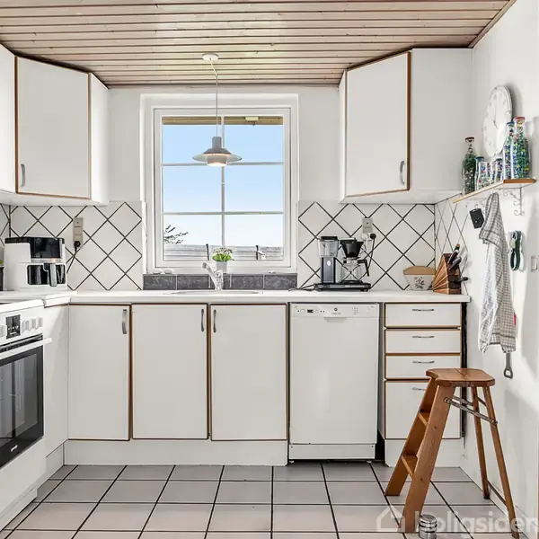 Køkken med hvidlakerede skabe og sorte håndtag, står stille, med en moderniseret ovn og opvaskemaskine. En trælåge til højre og vindue udsigt til have i baggrunden.