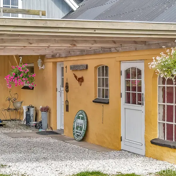 Et gult murstenshus med en overbygget terrasse. Dekoreret med blomster i hængepotter og en cirkulær metafacetavle. Tekst: "Skibsdrengen". Grusbelagt gårdsplads omgivet af grønne områder.
