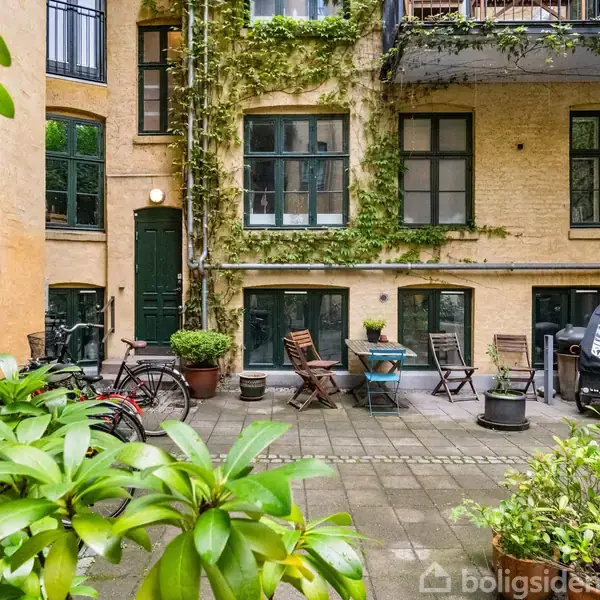 En hyggelig gårdhave med fliser omgivet af en gul murstensbygning med klatreplanter. Der er borde, stole og cykler placeret omkring gårdhaven, der er rig på grønne planter.