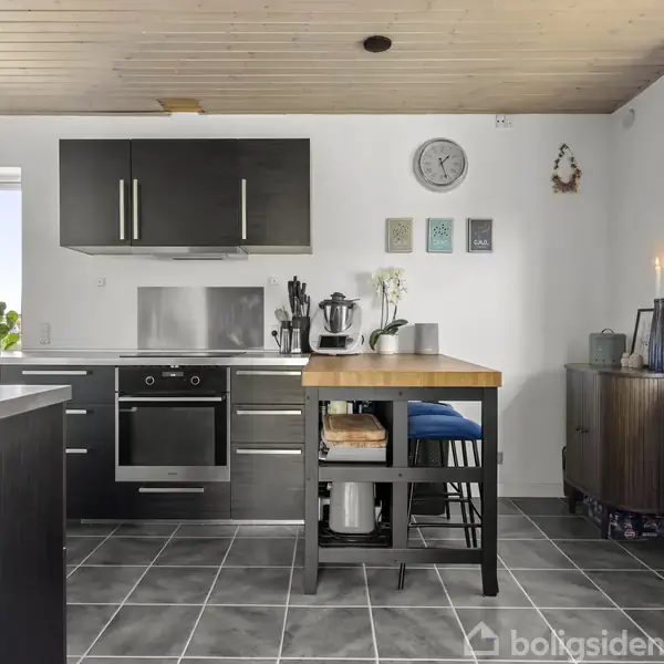 Stue med lyse træmøbler, en blå sofa, og kunst på væggen. Store vinduer giver naturligt lys. Spisebord med forskellige stole i et lyst, rummeligt miljø.