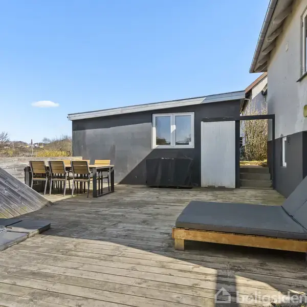 Solstol og spisebord står på en træterrasse, hvor der er et hegn mod en græsplæne og beplantning. Huset har hvide vægge med sorte detaljer under en blå himmel.