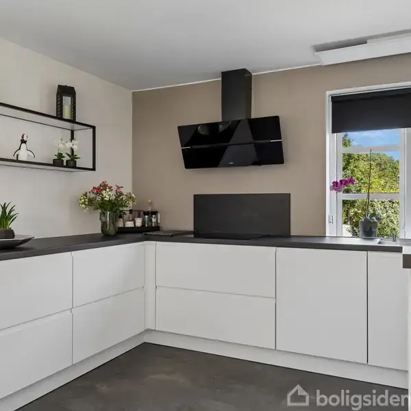 Moderne køkken med hvide skabe og mørke bordplader. En sort emhætte hænger over kogepladen. Der er en blomsterfyldt vindueskarm, væghylde med planter, og dekorationer.