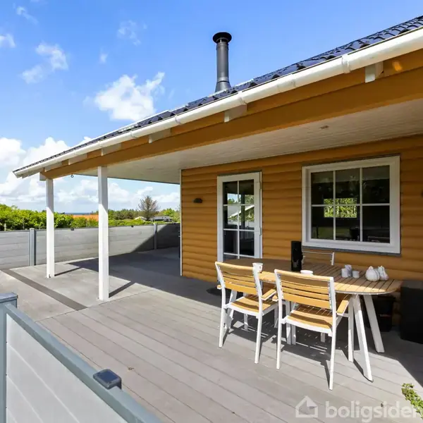 Træhus med terrasse; bord og stole står udenfor huset. Taget har en skorsten. Huset er omgivet af grønt landskab under en blå himmel med skyer.