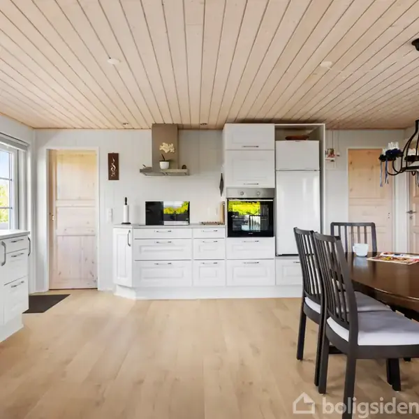 Et lyst køkken-alrum med hvidt køkkenindretning, indbygget ovn, sort spisebord med stole og stor lysekrone. Træloft og store vinduer giver naturligt lys, trægulve skaber en varm atmosfære.