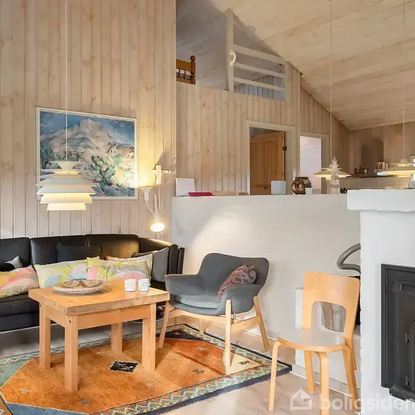 En sort sofa står i et hyggeligt stueområde med et træbord foran. En brændeovn til højre varmer rummet, som er lyst og indrettet med træpaneler og farverige tæpper.