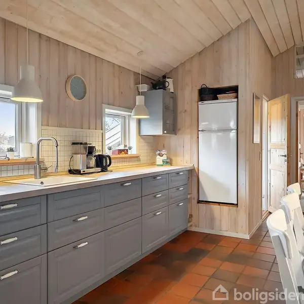 Et køkken med grå skabe under en lang bordplade. Der er to vinduer over vasken, og en lodret fryser ved væggen. Fliser på gulvet. Ingen tekst til stede.