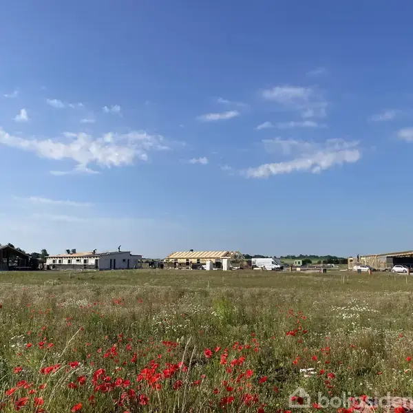 Byggeplads med flere huse under opførelse i det fjerne. Foran ses en mark med røde blomster, omgivet af åbent landskab og blå himmel med spredte skyer.