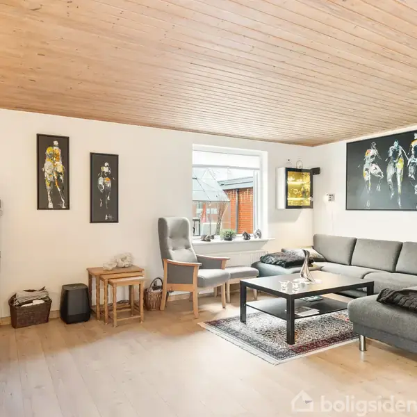 Gråt sofahjørne med sofabord, placeret i stue med trægulv og træloft. Vægge prydet med moderne kunst. Et vindue viser udsigt til nabobygningens facade.