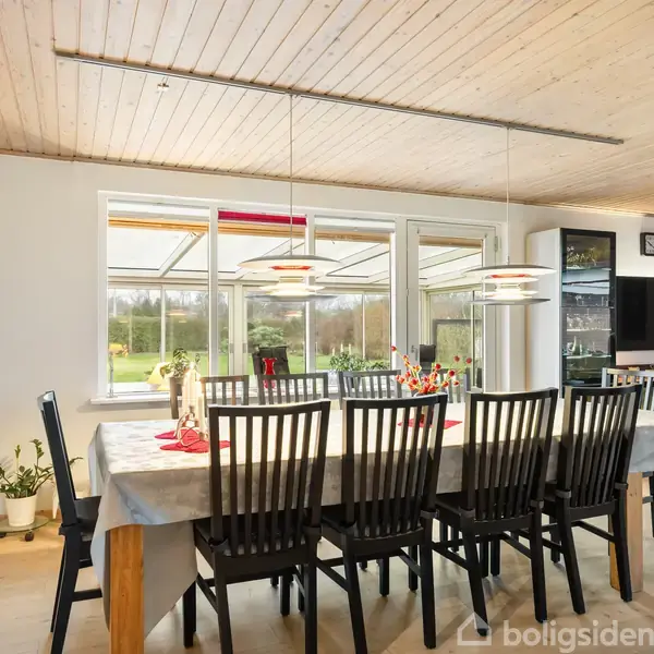 Langt spisebord med otte sorte stole står i en lys spisestue. Bordet er dækket med en lys dug. Store vinduer viser en grøn have udenfor.