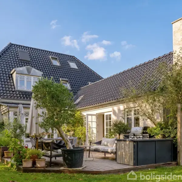 En hvid villa med mørkt tag står i sollys. En sort bil holder parkeret under en carport til venstre. Omkring huset er der grønne buske og træer.