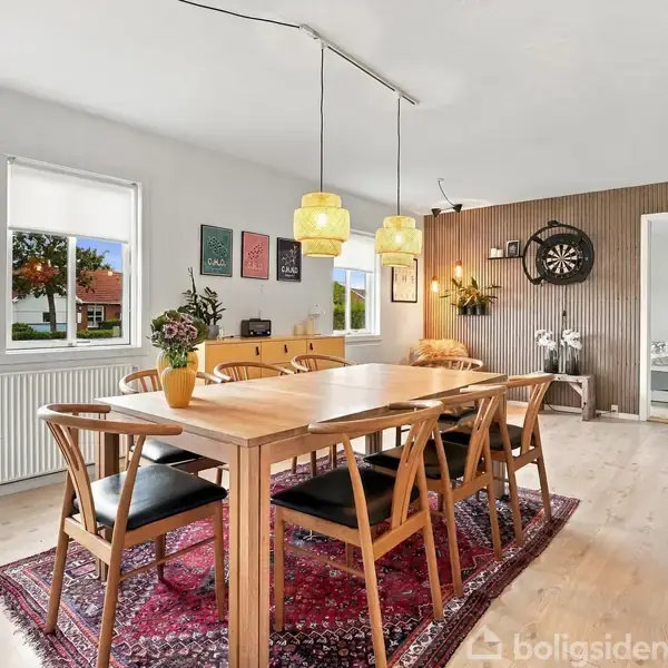 Et spisebord er omgivet af seks stole, placeret på et rødt tæppe. Over bordet hænger to gule lamper. Rummet har lyse trægulve, vinduer og en væg med kunst.