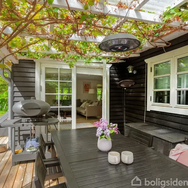 Sort træterrasse med spisebord og grill, omkranset af grønne planter på pergola. Åben dør viser kig ind til lys stue. Blomsterkrukke og stearinlys pynter bordet.