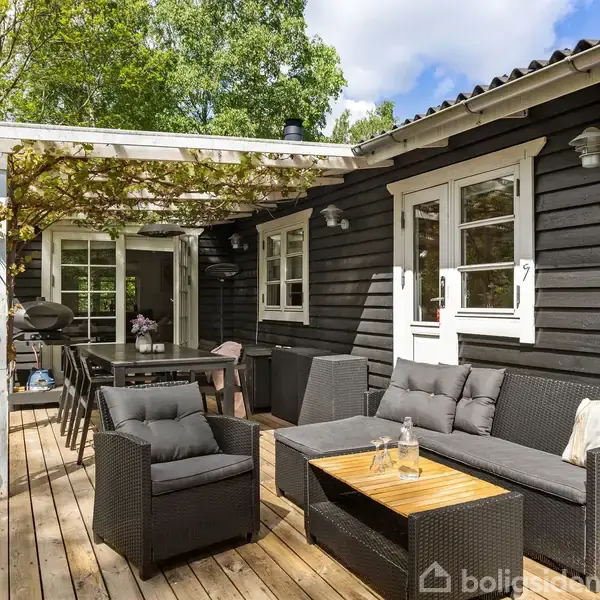 Sort træhus med hvidrammede vinduer, omgivet af grønne træer. Terrasse er pyntet med sorte kurvemøbler, et træbord og en pergola. I baggrunden ses blomsterdekorationsdetaljer.