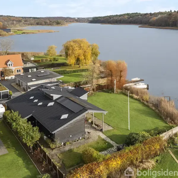 Flere huse står langs en sø med en stor have foran, omgivet af efterårsføjede træer. Søen strækker sig i baggrunden med skov på modsatte bred.