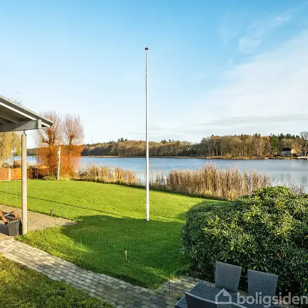 En sø med omgivende natur er synlig fra en terrasse med havemøbler, værende del af en bolig med græsplæne. himlen er blå, og vejret ser klart og solrigt ud.
