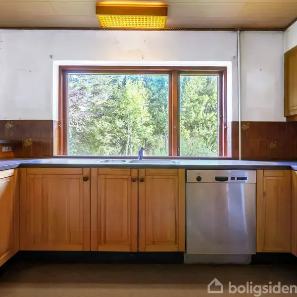 Køkken med træskabe og indbygget opvaskemaskine står stille. I baggrunden ses vinduer med udsigt til grønne træer. Lysebrun bordplade og hvid loftsbelysning fuldender indretningen.