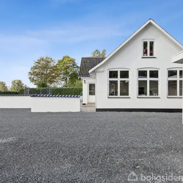 Hvidt hus med store vinduer står stille i en gårdsplads dækket af grus. Til højre ses en carport, mens træer og marker er synlige i baggrunden.