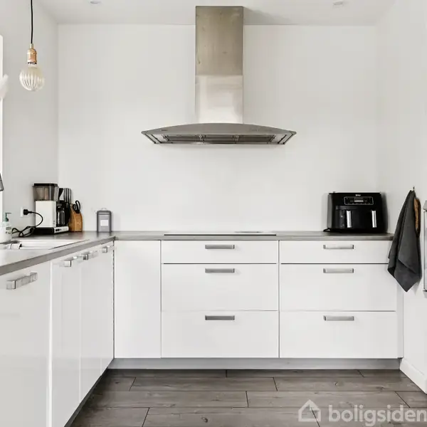 Et moderne køkken med hvide skabe og en stål-udsugning. En airfryer står på bordet ved siden af ovnen; en kaffemaskine og brødrister står ved vinduet.