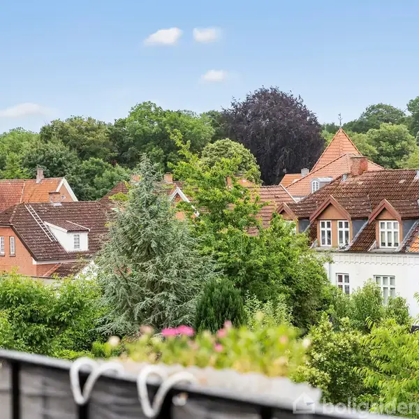 Røde murstenshuse med skrå tage omgivet af tætte, grønne træer og buske. Skyfri blå himmel danner baggrunden. En altan eller terrasse med en blomsterkasse i forgrunden.
