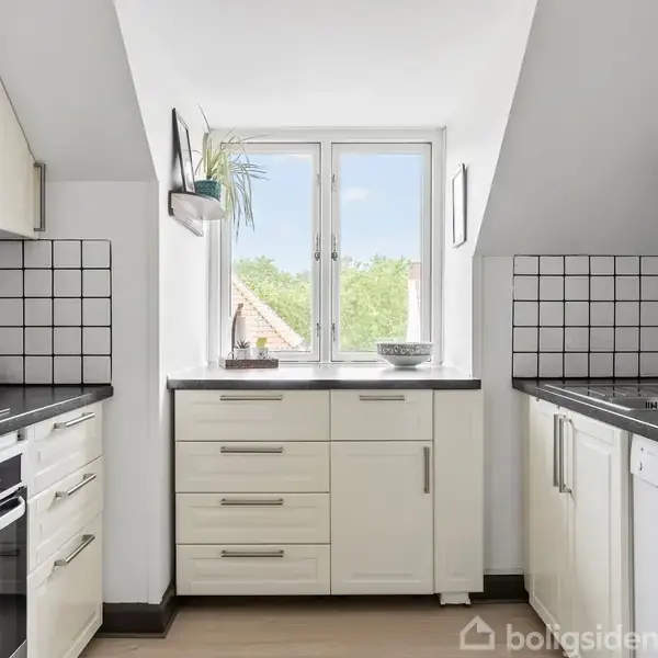 Et moderne køkken med hvide skabe og sorte bordplader. Øverst ses en ovn, håndvask, og knivblok. Vinduet giver naturligt lys med udsigt til træer. Tilede vægge tilføjer et klassisk touch.