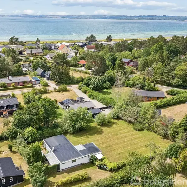 Huse ligger spredt i et frodigt grønt område tæt ved kysten. Nogle huse har store græsplæner, og baggrunden viser havet og en skovklædt kystlinje under en blå himmel.