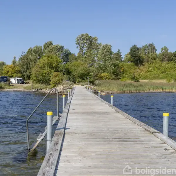 En træbro strækker sig over en sø mod en frodig skov. Vandet er roligt, og himlen er klar blå. En bil og campingvogne ses i baggrunden mellem træerne.