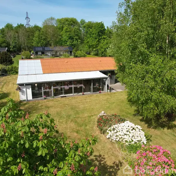 Hus med orange tag står på en grøn græsplæne omgivet af blomsterbede og høje træer. I baggrunden ses flere huse blandt frodig vegetation under en klar blå himmel.