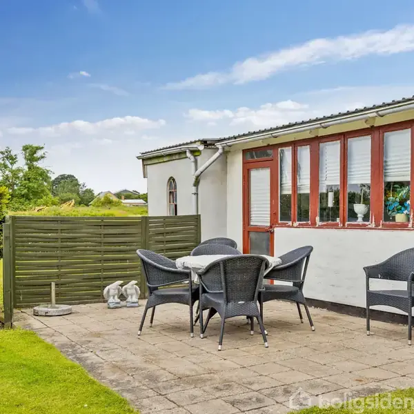 En udendørs terrasse med et rundt bord og fem mørke stole. Terrassen er flisebelagt og støder op til en hvid bygning med røde vinduer. Der er stillet skulpturer på græsplænen.