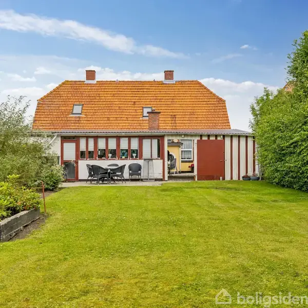 Hus med rødt tegltag og hvid facade står i en frodig have med grøn plæne. Række af havestole og bord på terrasse foran huset.