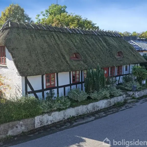 Bindingsværkshus med stråtag står langs en stille vej, omgivet af grønne buske og træer. Flere små vinduer og en rød dør ses på husets facade.