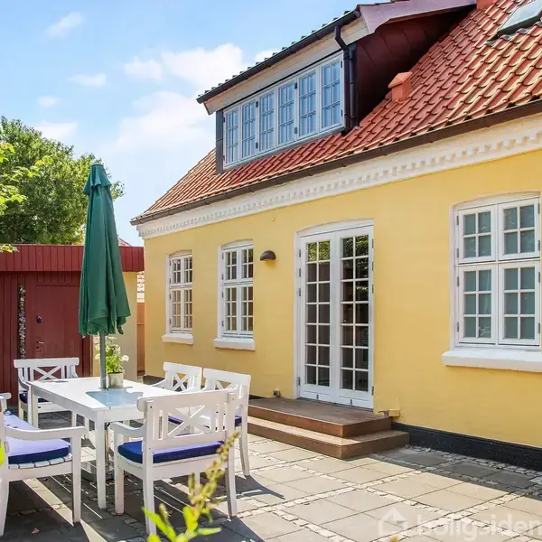 Gul husfacade med rødt tegltag. Et havesæt med parasol står foran huset på en flisebelagt terrasse. Grønt hegn og planter omkranser området under en blå himmel.