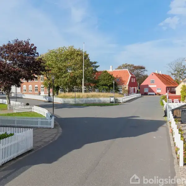 En tom gade strækker sig forbi farverige huse med røde tage. Hvide stakitter omkranser haverne. Træer står ved vejen, og himlen er blå med spredte skyer.