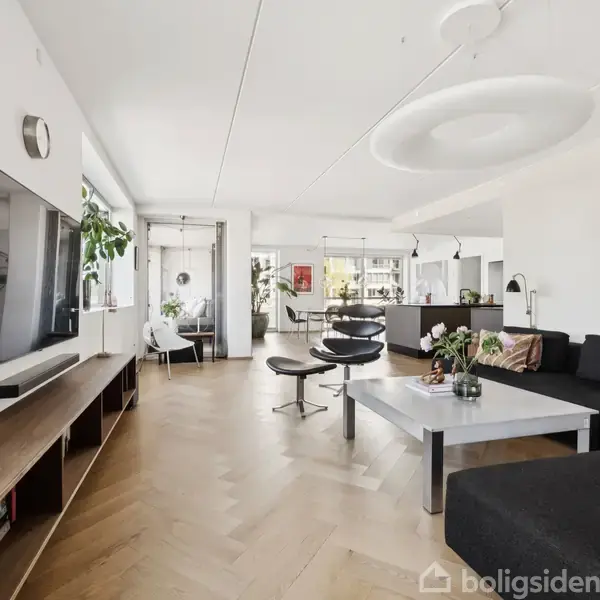 Stue med sort sofa og fladskærms-TV på væggen, moderne indretning og træmøbler. Store vinduer slipper lys ind i rummet, der er dekoreret med kunst og planter.