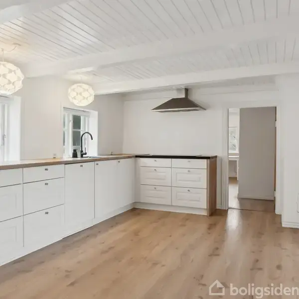Hvidt køkken med træbordplader og en sort emhætte i et lyst rum. Store vinduer lukker naturligt lys ind. Lyse trægulve og hvide vægge dominerer rummet, hvilket skaber en modernistisk stil.