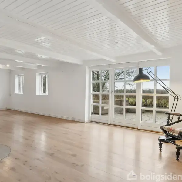 Stue med en gyngestol og en lampe i hjørnet. Store vinduer åbner ud mod en terrasse med udsigt til natur. Trægulv og hvidmalet træloft skaber en lys atmosfære.